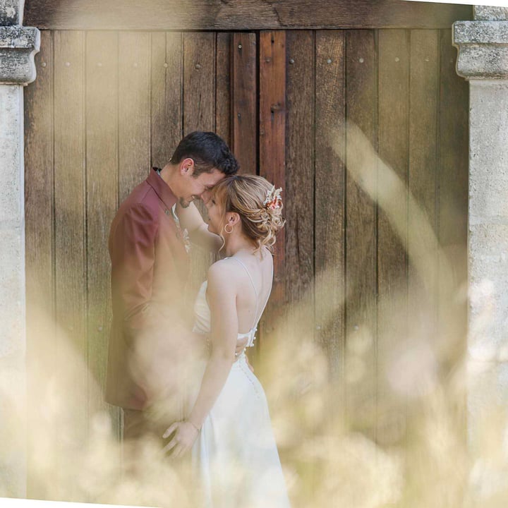 Un couple de jeunes mariés se tenant tendrement devant une porte en bois, entourés de flou artistique créé par des herbes hautes, symbolisant l'amour et l'intimité.
