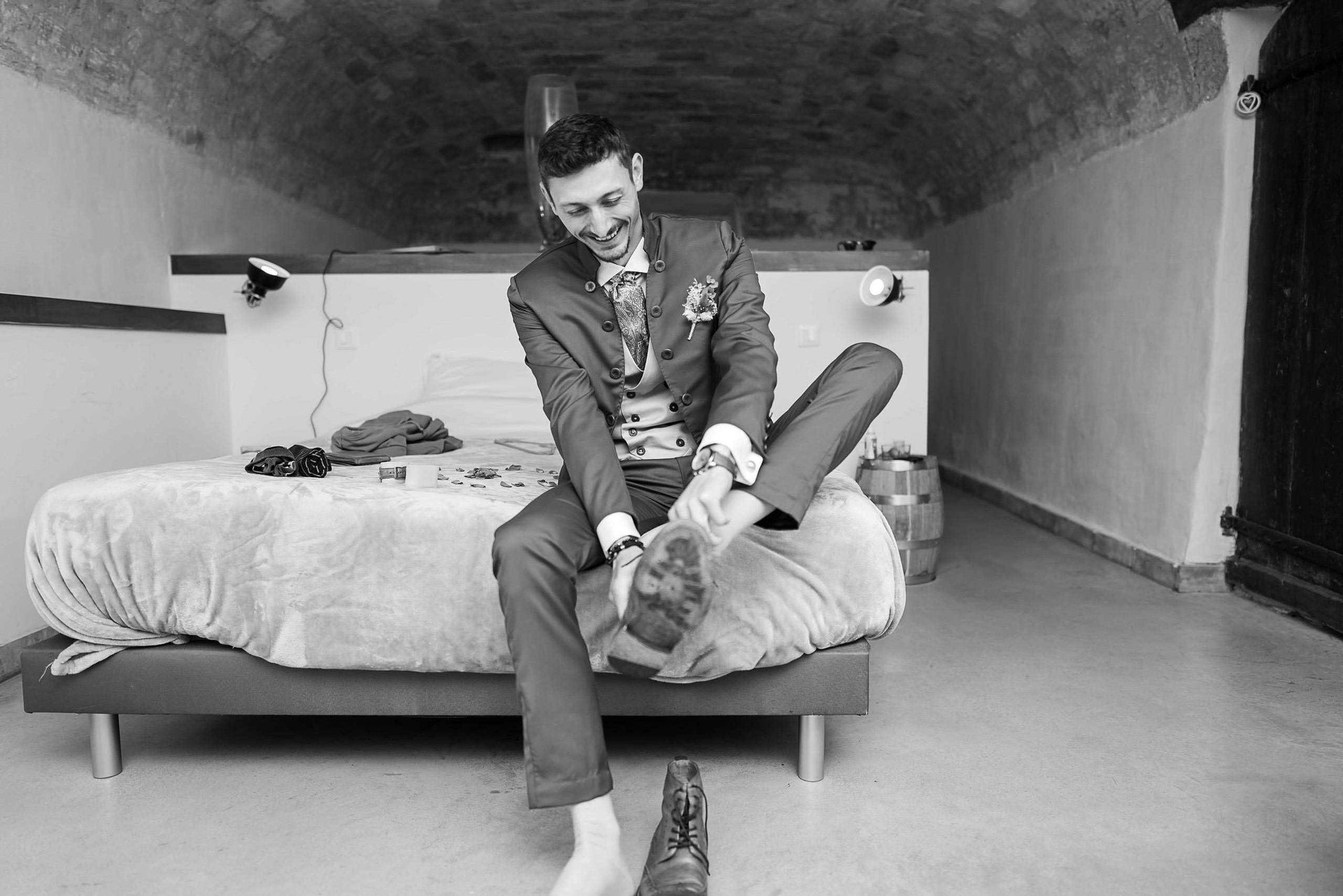 Un homme en costume assis sur un lit, souriant, en train de mettre ses chaussures, avec des vêtements éparpillés sur le lit et un tonneau en bois visible en arrière-plan.