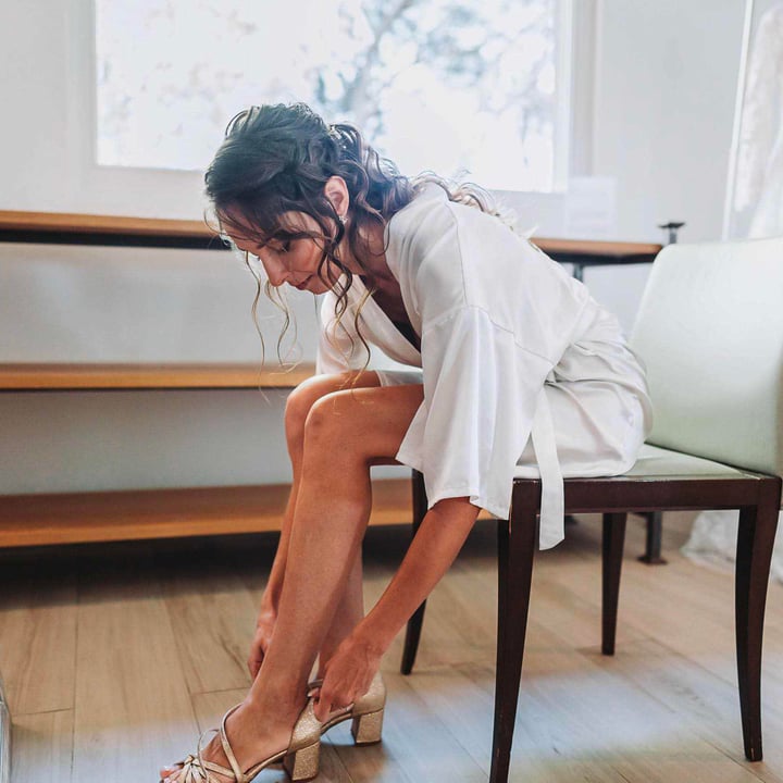 Une mariée assise sur une chaise, en train de mettre ses chaussures à talons dorés, vêtue d'un peignoir blanc, avec une robe de mariée accrochée en arrière-plan.