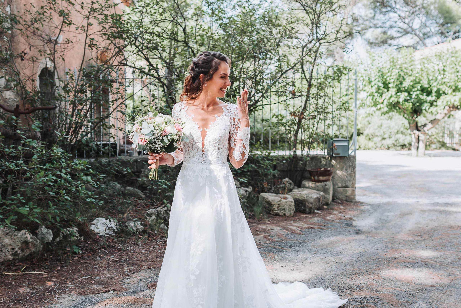 Une mariée souriante porte une robe de mariée à manches longues en dentelle, tenant un bouquet de fleurs dans sa main, sur un chemin entouré de verdure et de pierres.